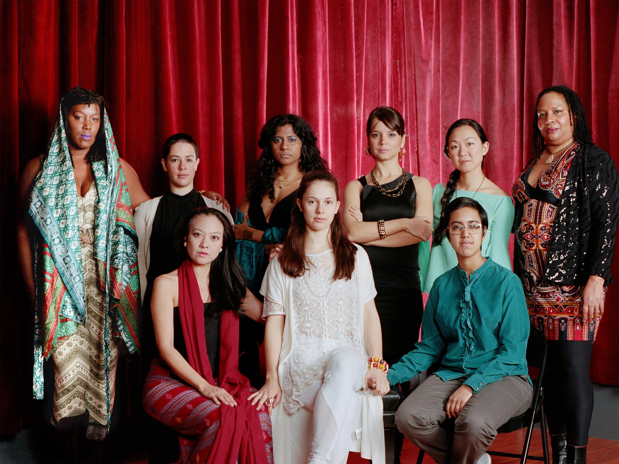 Group portrait of nine women posed in front of deep red stage curtains, wearing a mix of elegant, colorful, and patterned clothing, with some seated and others standing, all facing the camera with composed, serious expressions.