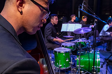 A close-up of a musician playing a double bass, with a drummer at a green drum kit and other ensemble members visible on stage in the background under concert lighting.