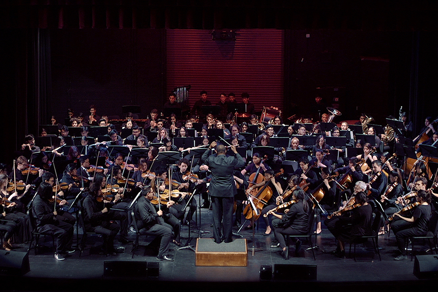 A large orchestra performs on a darkened stage, with a conductor standing at the center and musicians seated in sections of strings, woodwinds, brass, and percussion under dramatic stage lighting.