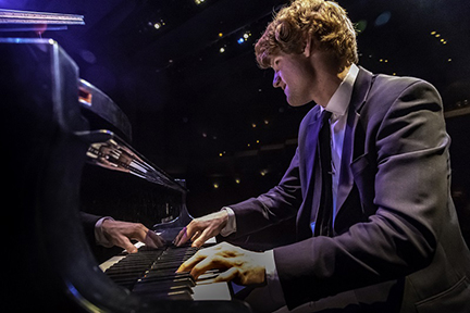 A pianist in a suit plays a grand piano on stage, his hands on the keys and body leaning in with concentration, as stage lights and reflections illuminate the instrument.