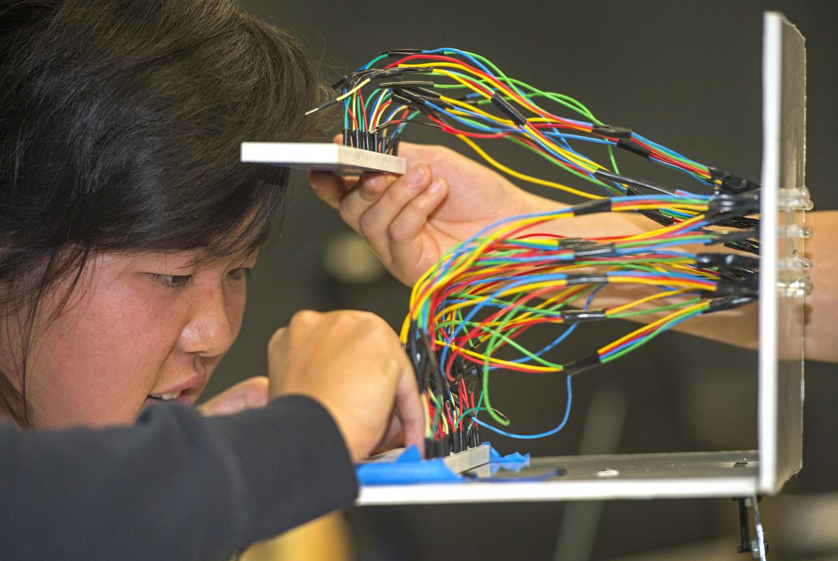A person concentrates while working on an electronic setup, holding a bundle of multicolored wires in one hand and making adjustments to components mounted on a metal frame.