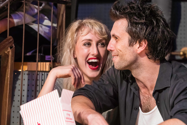 Two actors sit close together on stage, smiling and laughing, with one holding a striped popcorn container as they lean toward each other in a playful, intimate moment under warm theatrical lighting.