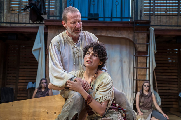 Two actors perform on a stage set resembling a rustic interior, with a man standing behind and supporting a distressed woman who clutches his arm, while other performers watch from the background under theatrical lighting.