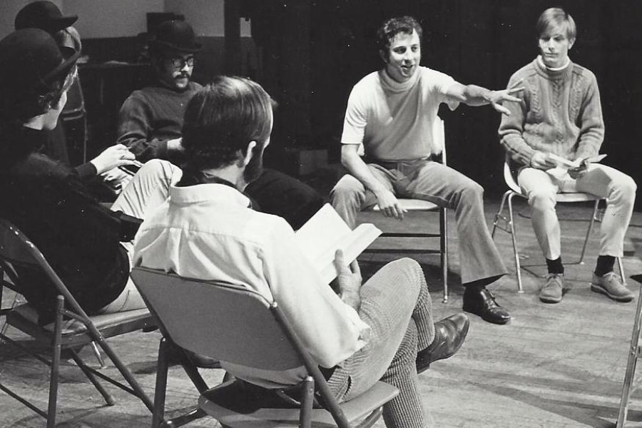 A black-and-white photo shows a group of people seated in a circle on folding chairs, engaged in discussion, with one person gesturing while others listen or read from papers in a rehearsal or workshop setting.
