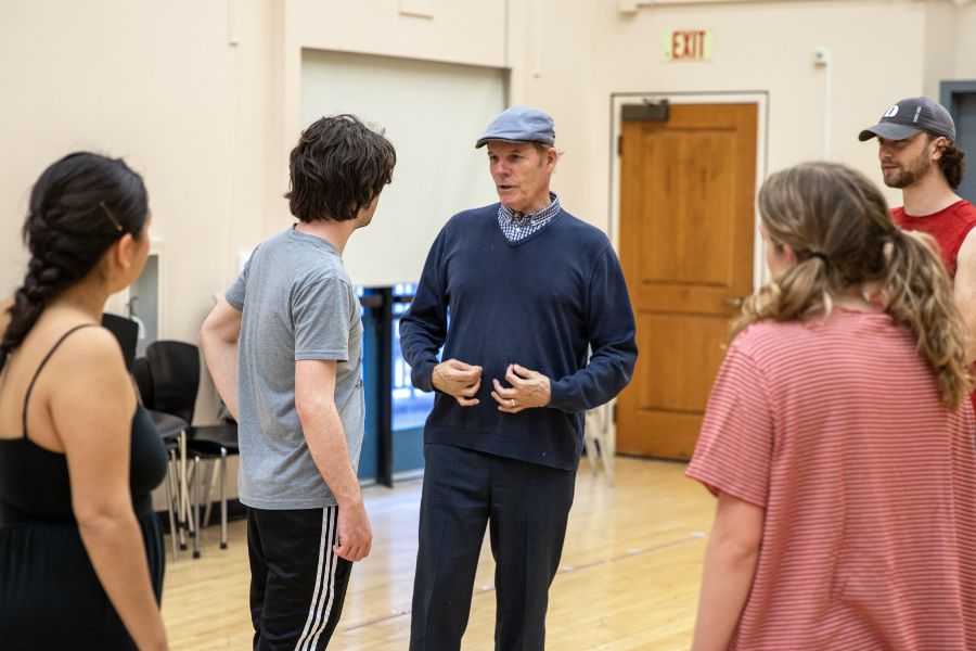 A group of people stand in a rehearsal studio, listening as a man wearing a cap and sweater speaks and gestures, with chairs and a door visible in the background.