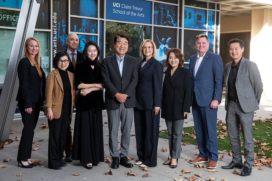 A group of adults stand together outside the UCI Claire Trevor School of the Arts, smiling and posing for a photo in front of glass windows and signage, dressed in professional attire with fallen leaves scattered on the ground.