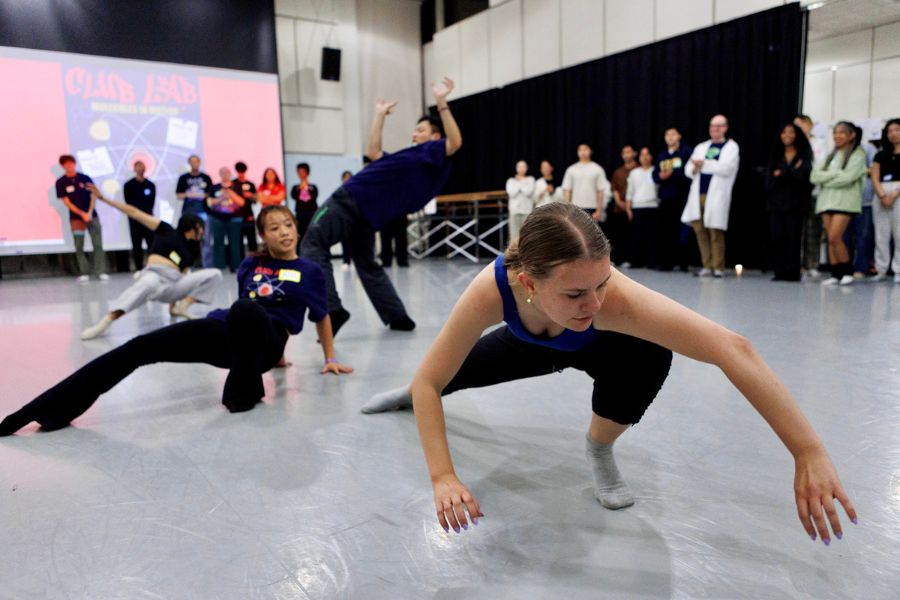 Dancers rehearse in a studio, with one dancer in the foreground reaching low toward the floor while others move dynamically behind her, as a group of people watch from the edges of the room.