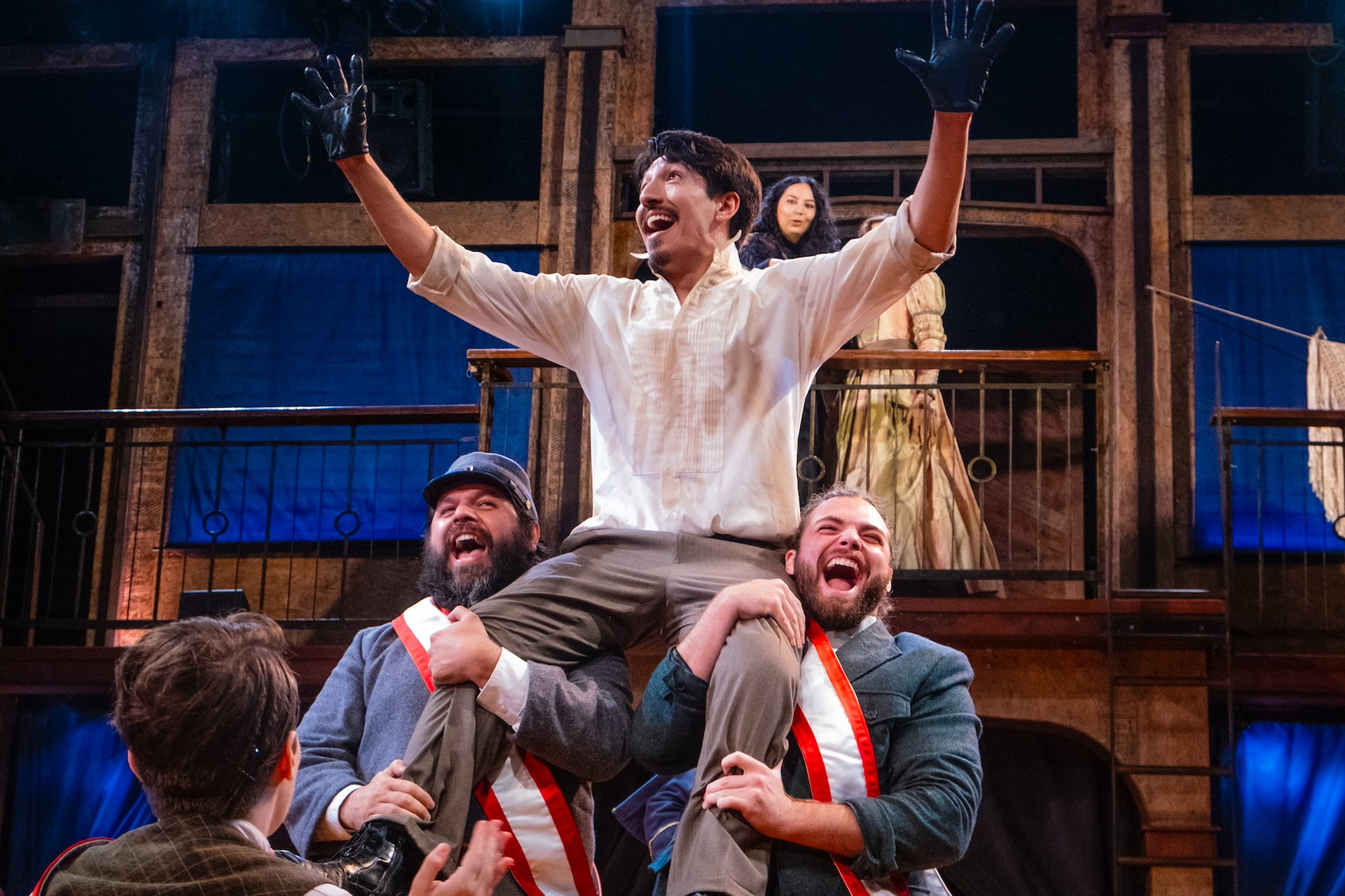 A theatrical scene shows a jubilant man lifted on the shoulders of two others, arms raised and wearing gloves, while additional actors stand and watch from a wooden set behind them under stage lighting.