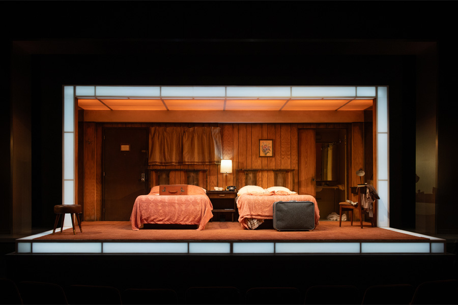 A stage set depicts a mid-century style bedroom with two twin beds, wooden walls, a bedside lamp, and period furniture, framed as if viewed through a large rectangular opening.