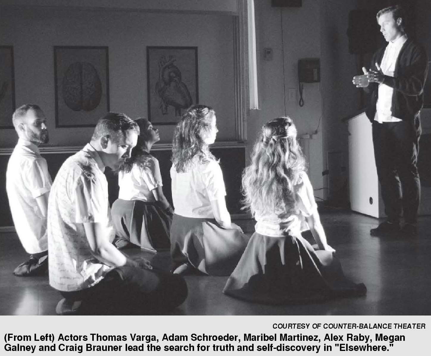 A black-and-white stage photograph shows several actors kneeling on the floor facing a standing speaker who addresses them, suggesting a theatrical rehearsal or performance scene set in a sparse room.