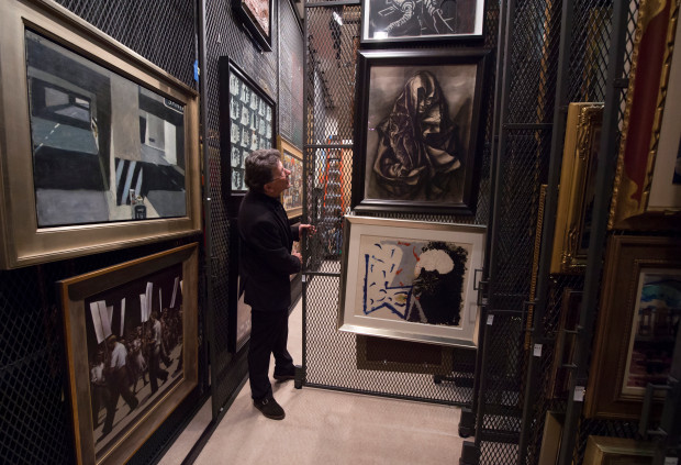 Dean Stephen Barker stands in a narrow, dimly lit art storage corridor, surrounded by large framed artworks stacked on rolling mesh racks. He looks closely at a display of black-and-white and monochrome pieces hung at varying heights, with tightly packed frames lining both sides of the aisle, creating a dense, archive-like museum environment.