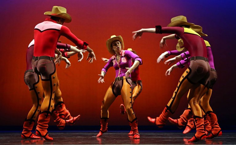 A group of dancers in coordinated cowboy-inspired costumes perform on stage, wearing wide-brimmed hats, fitted tops, chaps, and boots. They form a loose circle around a central dancer, all holding their arms bent and extended in stylized poses, against a vivid red-to-purple gradient backdrop that emphasizes the theatrical, high-energy choreography.