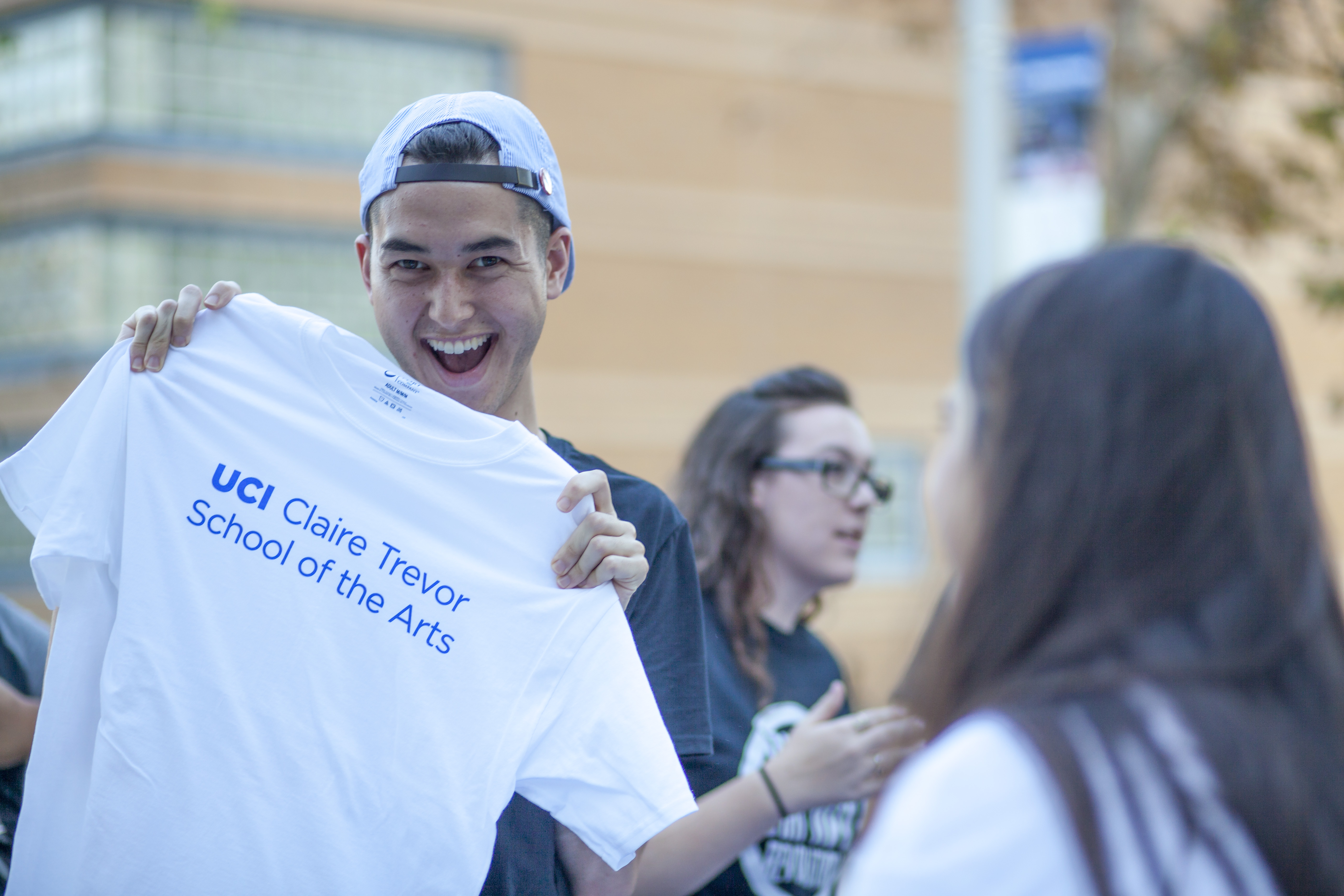 A smiling student holds up a white T-shirt that reads “UCI Claire Trevor School of the Arts,” presenting it enthusiastically to someone standing in front of him. He wears a backward baseball cap, while other students chat in the softly blurred background, suggesting a lively campus or arts-related event.