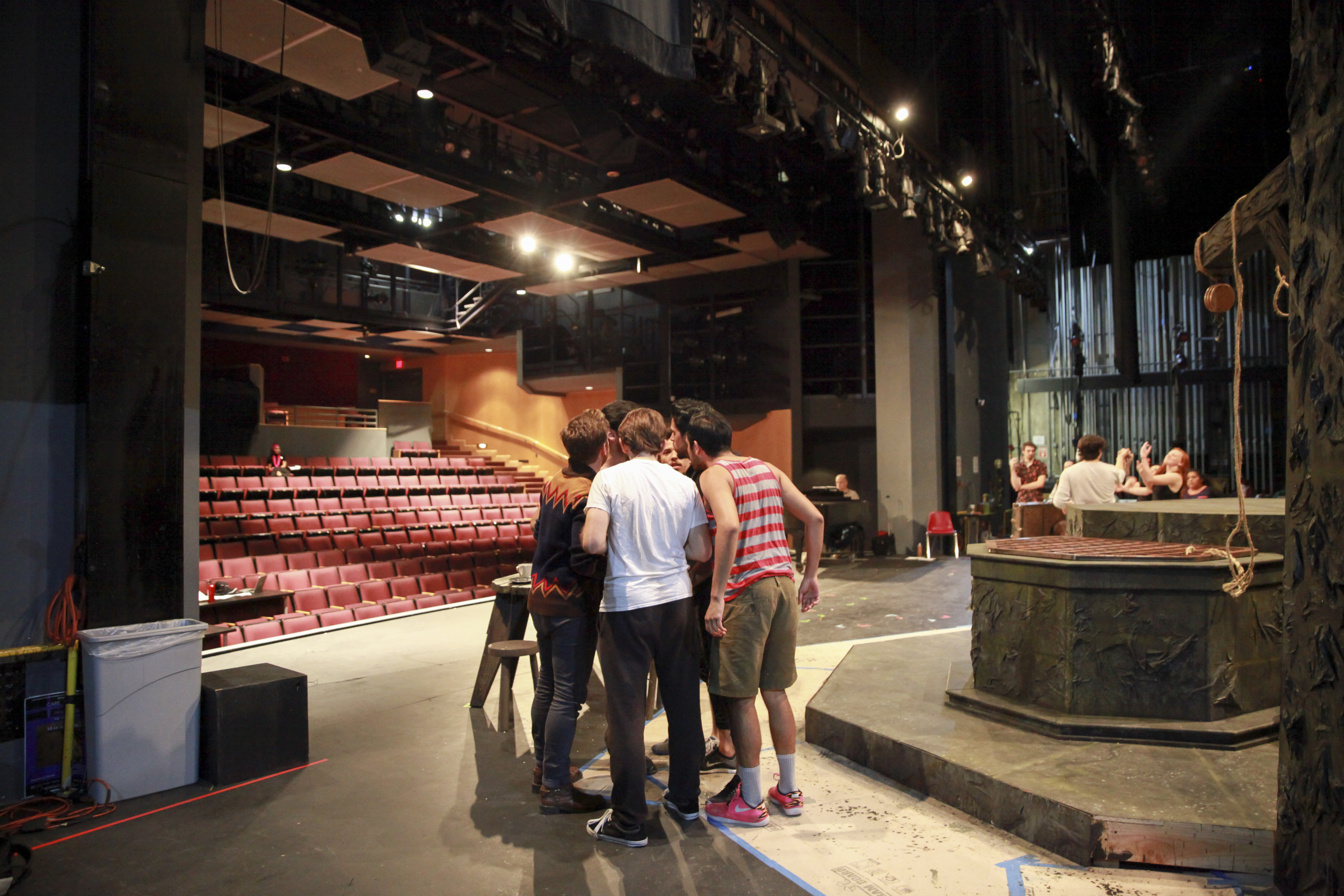 A group of people huddle together on a theater stage during rehearsal, standing near a set piece while talking closely. Rows of red audience seats stretch into the background, and stage lights, rigging, and curtains frame the space, offering a behind-the-scenes view of a production in progress.