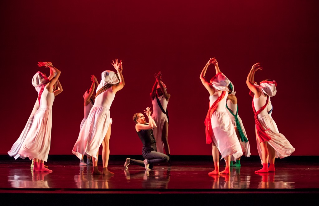 A group of dancers performs on stage in flowing white and red costumes, their arms lifted and curved overhead in synchronized, expressive shapes. One dancer kneels at the center while others form a loose semicircle around them, set against a deep red backdrop and reflected on the glossy stage floor, creating a sense of ritual and unity.
