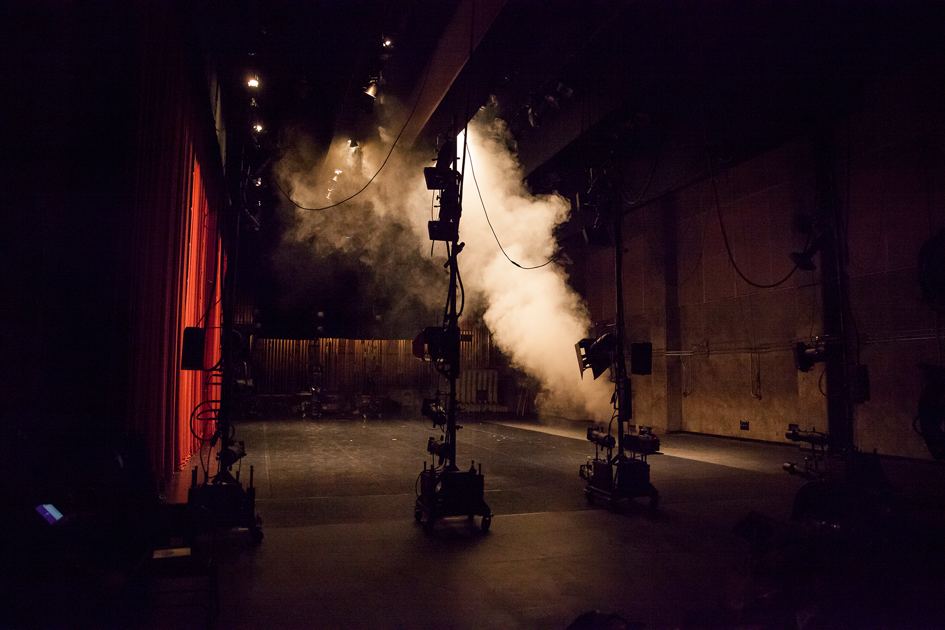 A dimly lit theater stage is shown from backstage, with lighting equipment and stands positioned across the floor as thick white fog drifts upward into the air. Red curtains line one side of the space, and the dark, industrial setting creates a moody, behind-the-scenes atmosphere just before or after a performance.