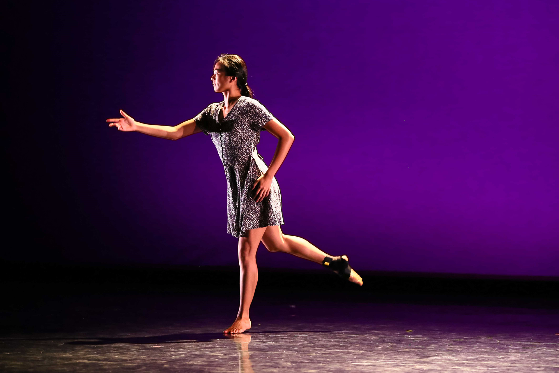 A solo dancer performs on a dark stage against a rich purple backdrop, standing on one leg with the other lifted behind her. She reaches one arm forward in a poised, expressive gesture, wearing a patterned short dress and bare feet, with the stage lighting highlighting her balance and controlled movement.