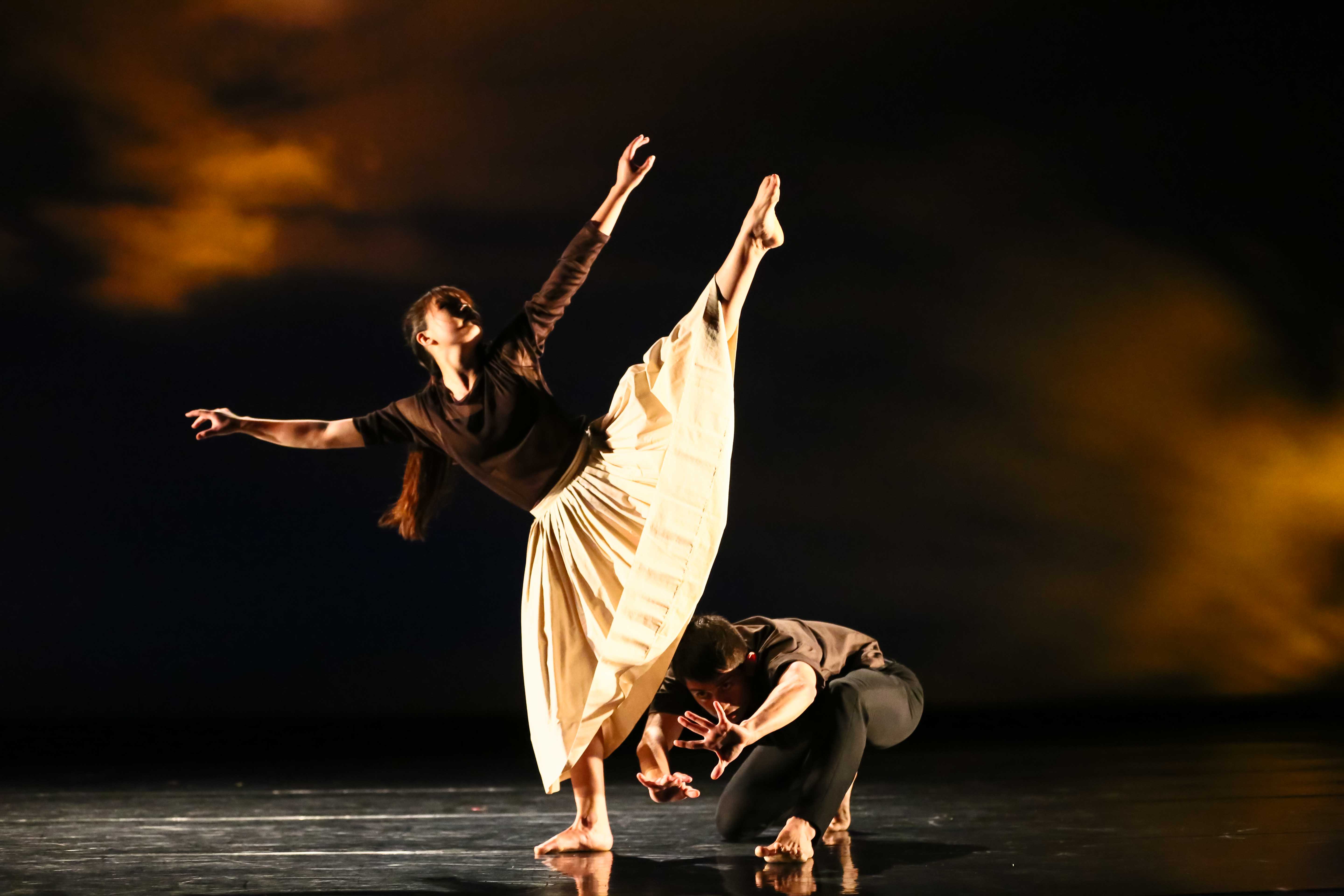 Two dancers perform on a dimly lit stage, one balancing on a single leg with the other leg extended high behind her and arms reaching outward in a controlled, sculptural pose. The second dancer crouches low near her feet, supporting or interacting with the movement, while warm, atmospheric lighting creates a dramatic contrast against the dark background.