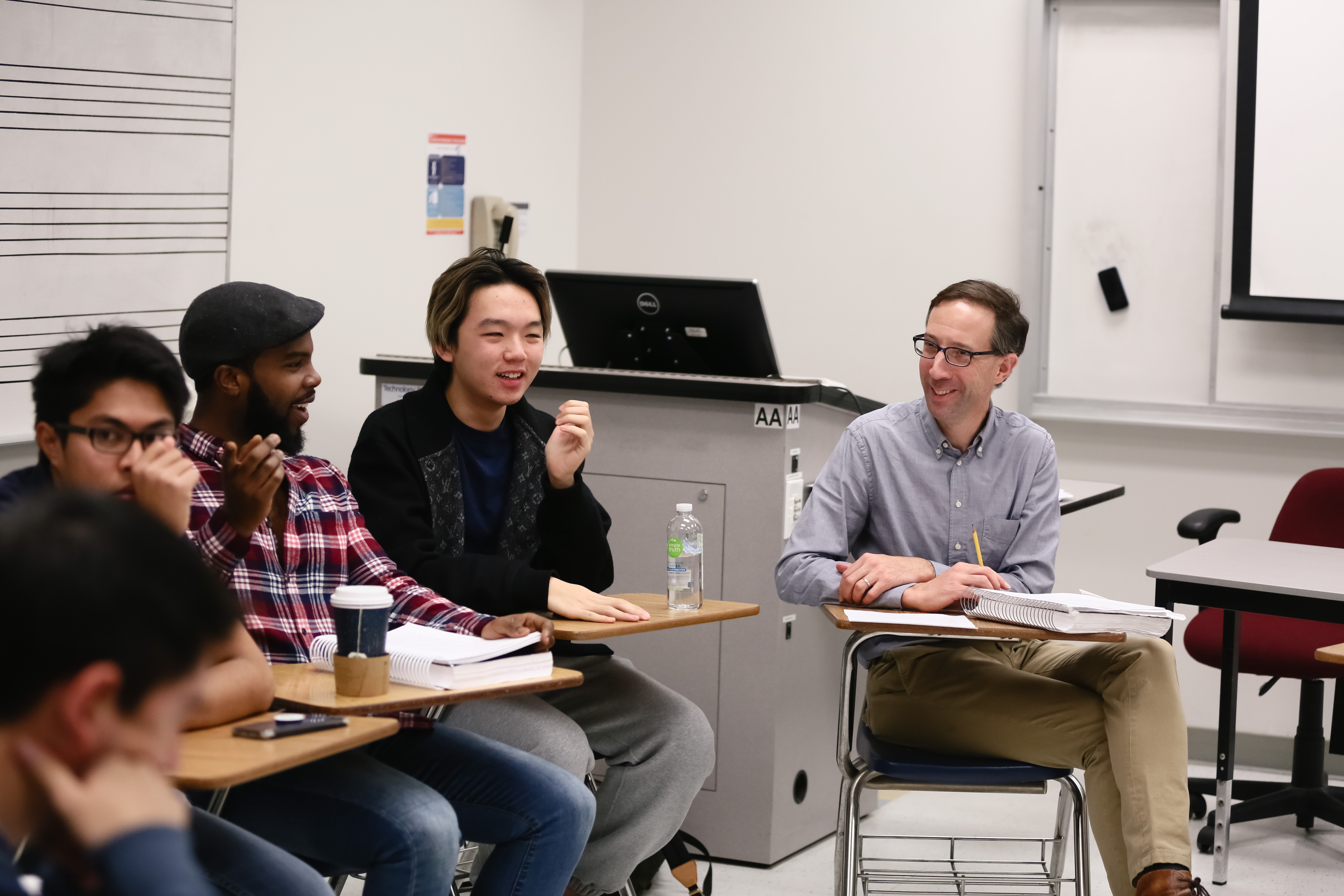Several people sit at desks in a classroom, facing one another in discussion. One person gestures with their hand while speaking, and others smile or listen attentively. Laptops, notebooks, and cups rest on the desks, with a whiteboard and podium visible in the background.