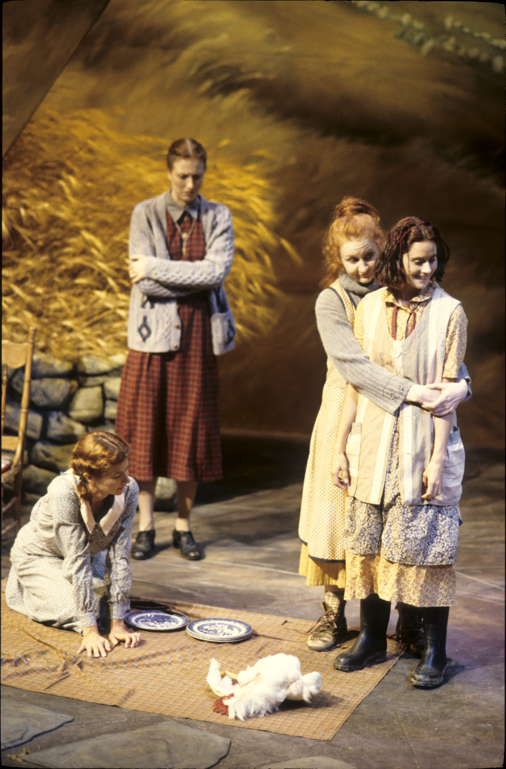 Four young women in period-style clothing stand on a stage with a warm, golden backdrop. Two stand closely together at center, one with her arms around the other, both facing forward. Another woman stands behind them with her arms crossed, watching, while a fourth kneels on the floor to the left, leaning on one hand near two plates. A small white chicken stands on a woven mat in the foreground. The lighting is theatrical, casting soft shadows across the wooden floor and stone elements behind them.