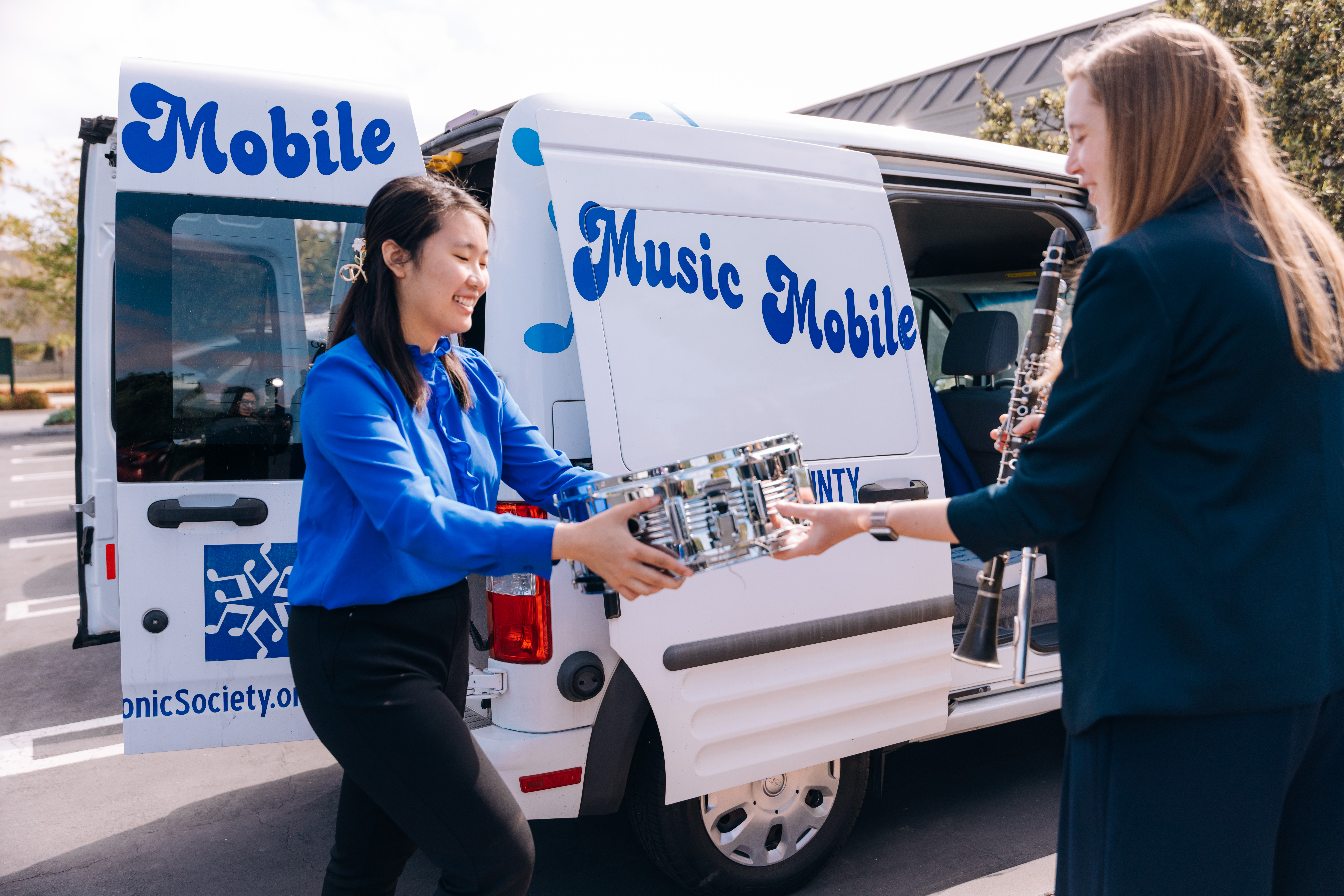 Two people stand beside a white van labeled “Music Mobile,” smiling as they exchange a small electronic device. One person holds a clarinet while reaching out, and the open van door reveals equipment inside, suggesting a music outreach or community program taking place outdoors in daylight.