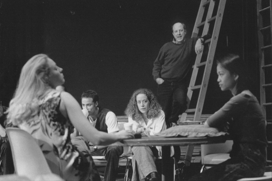 Several people gather around a table on a stage in a black-and-white scene. Four sit facing one another, leaning in with attentive or serious expressions, while one person stands behind them near a ladder, resting an arm on it as they observe. Chairs, the table, and minimal stage elements are visible against a dark background.