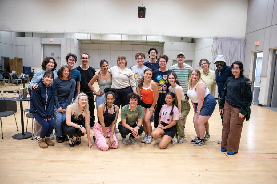 A large group of people pose together in a rehearsal studio, arranged in rows with some standing and others kneeling in front. They smile toward the camera, wearing casual clothing. The room has a wooden floor, mirrors along the back wall, and chairs and tables visible at the sides.