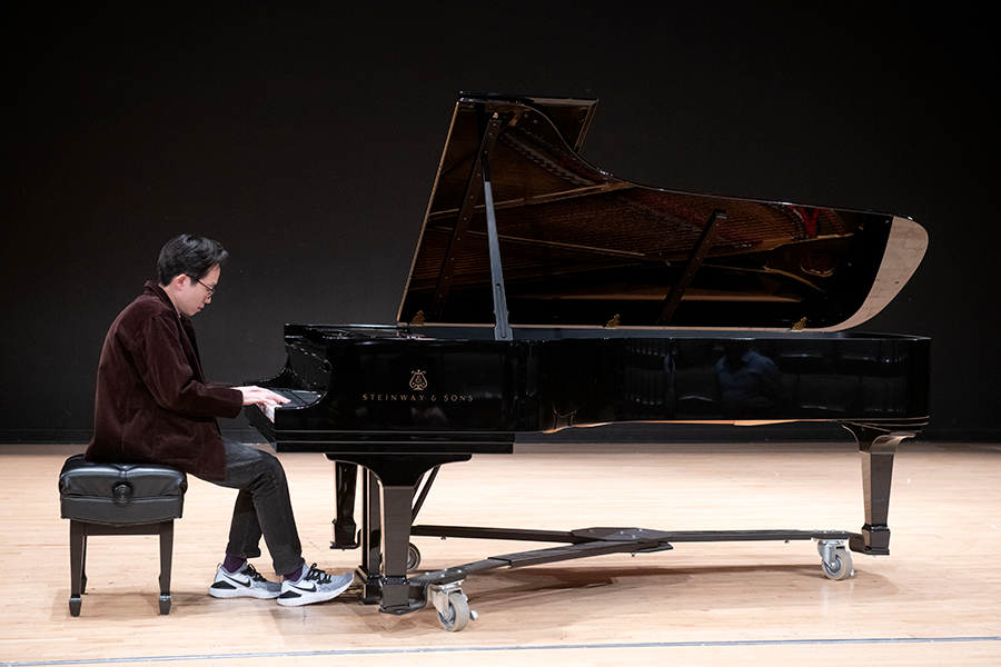 A pianist sits on a bench at a grand piano on a bare stage, playing with focused posture. The piano lid is raised, and the instrument rests on wheeled supports. The performer wears dark clothing and sneakers, and the scene is lit evenly against a black background.