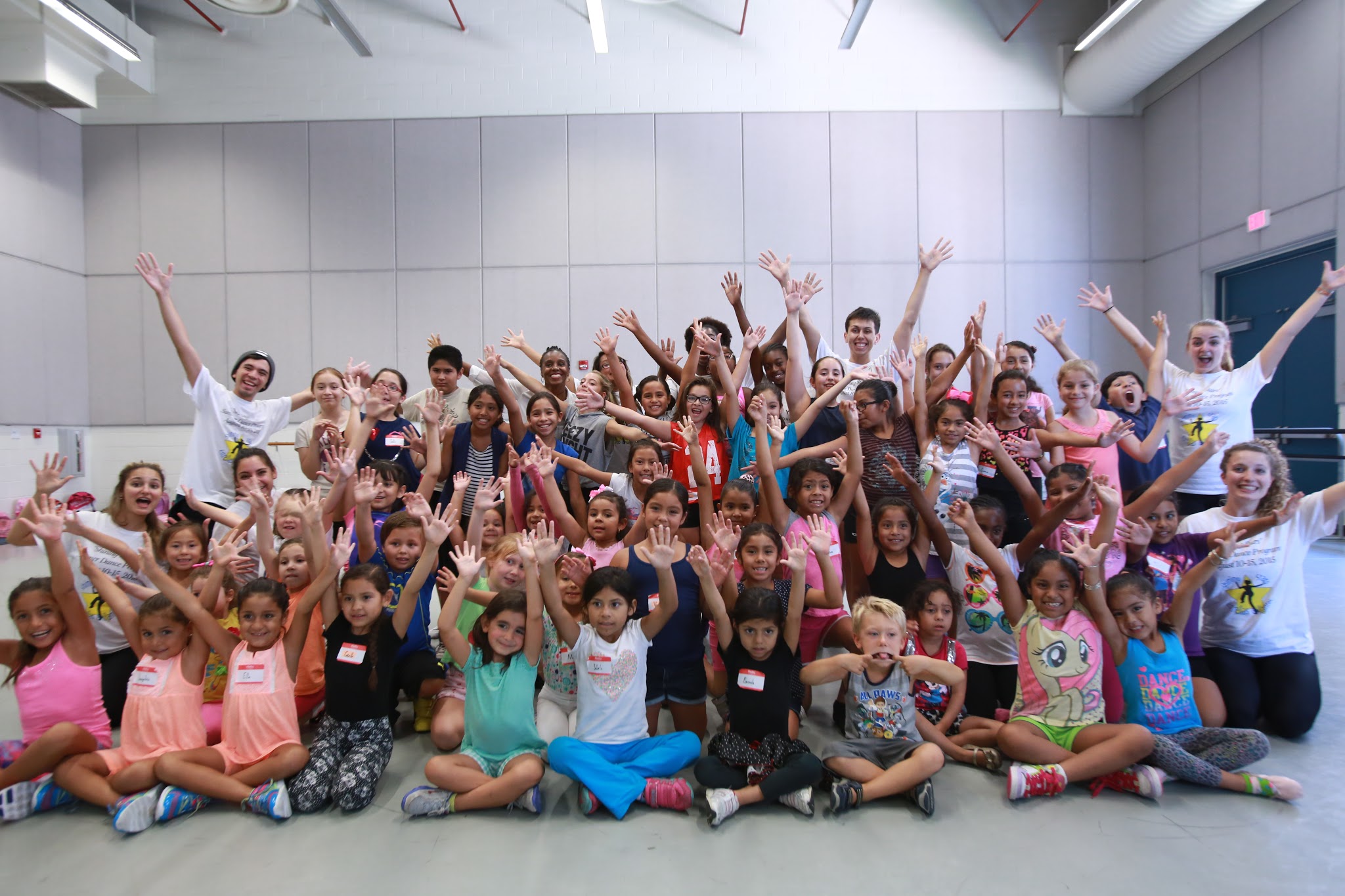 A large group of children and a few adults pose together indoors, seated and standing in rows. Many raise their hands and smile toward the camera. The room has light-colored walls and a high ceiling with exposed fixtures.