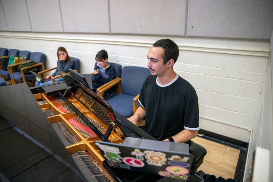 A person sits at a grand piano in a rehearsal room, reading from sheet music placed on the stand. Other individuals sit in chairs behind the piano, some looking down at their own materials, within a plain, well-lit studio space.