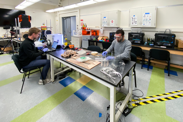 Two people work at a large table in a lab-like workspace, surrounded by tools, electronics, and equipment. One person uses a laptop while the other handles materials laid out on the table, with cabinets and workstations lining the room.