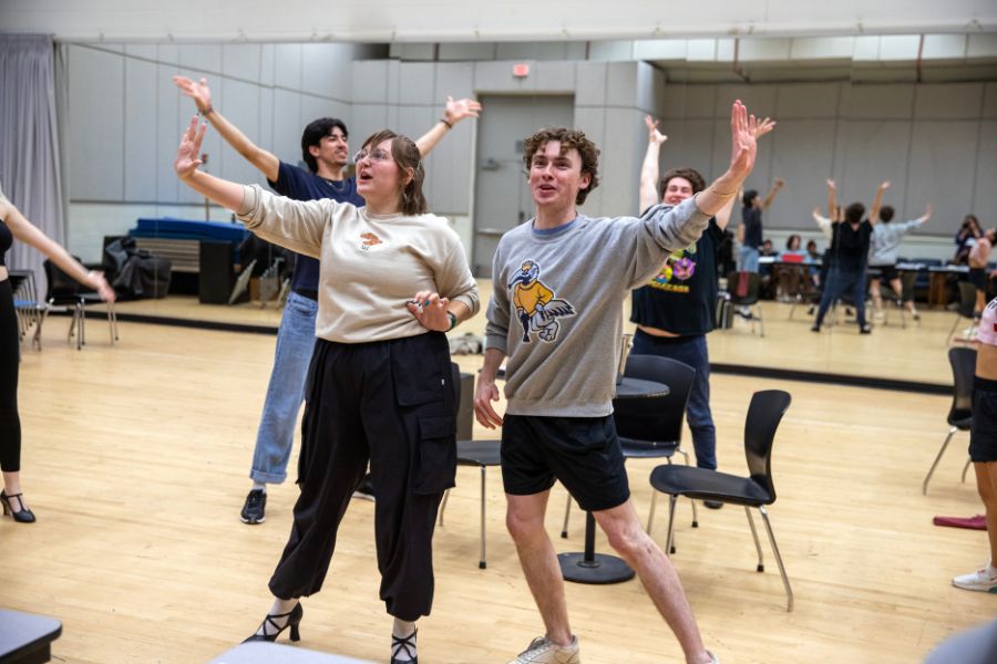 Several people stand in a rehearsal studio with arms raised in unison, arranged among scattered chairs. They wear casual clothing and face forward, captured mid-gesture within the bright, mirrored space.