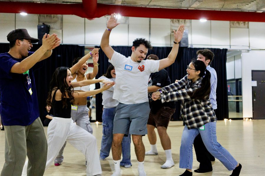 A group of people gathers in a studio space around one person at center with raised hands. Others reach toward them from all sides, creating a sense of collective motion and interaction within the room.