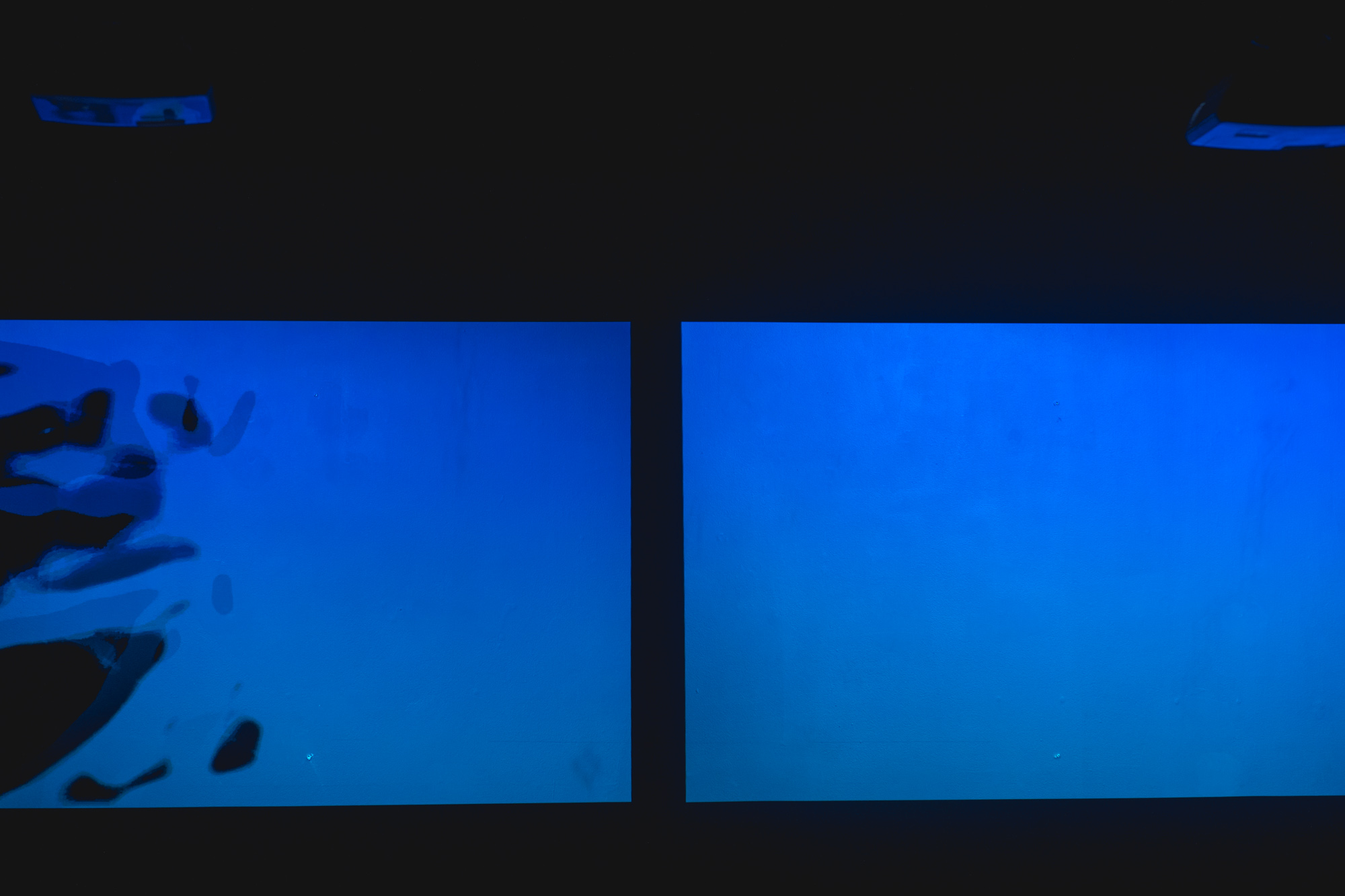 Two rectangular blue video projections appear side by side against a dark background. The left screen shows abstract, fluid shapes drifting across the blue field, while the right screen remains mostly uniform in color.