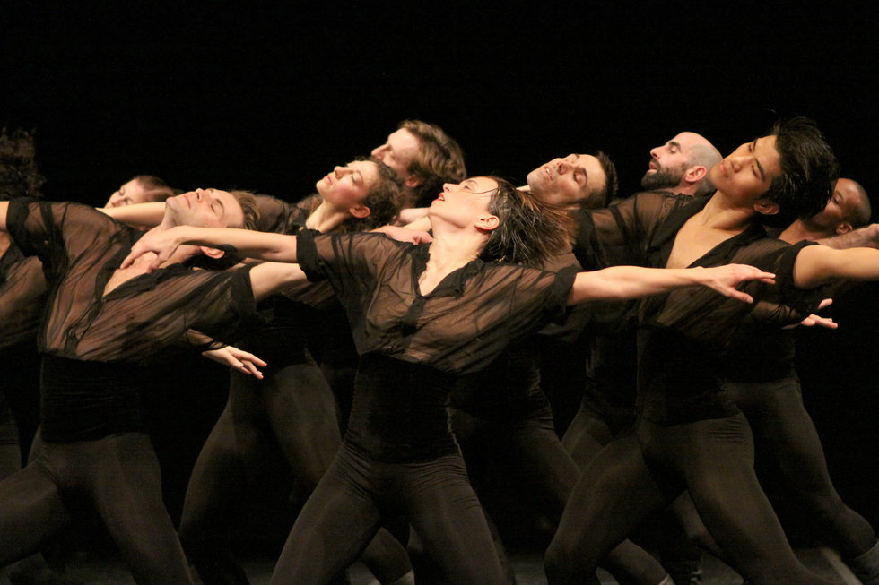 A group of dancers moves together in a tightly layered formation on a dark stage. Dressed in sheer black costumes, they lean and extend their arms in unison, creating a sweeping, interconnected shape under focused lighting.