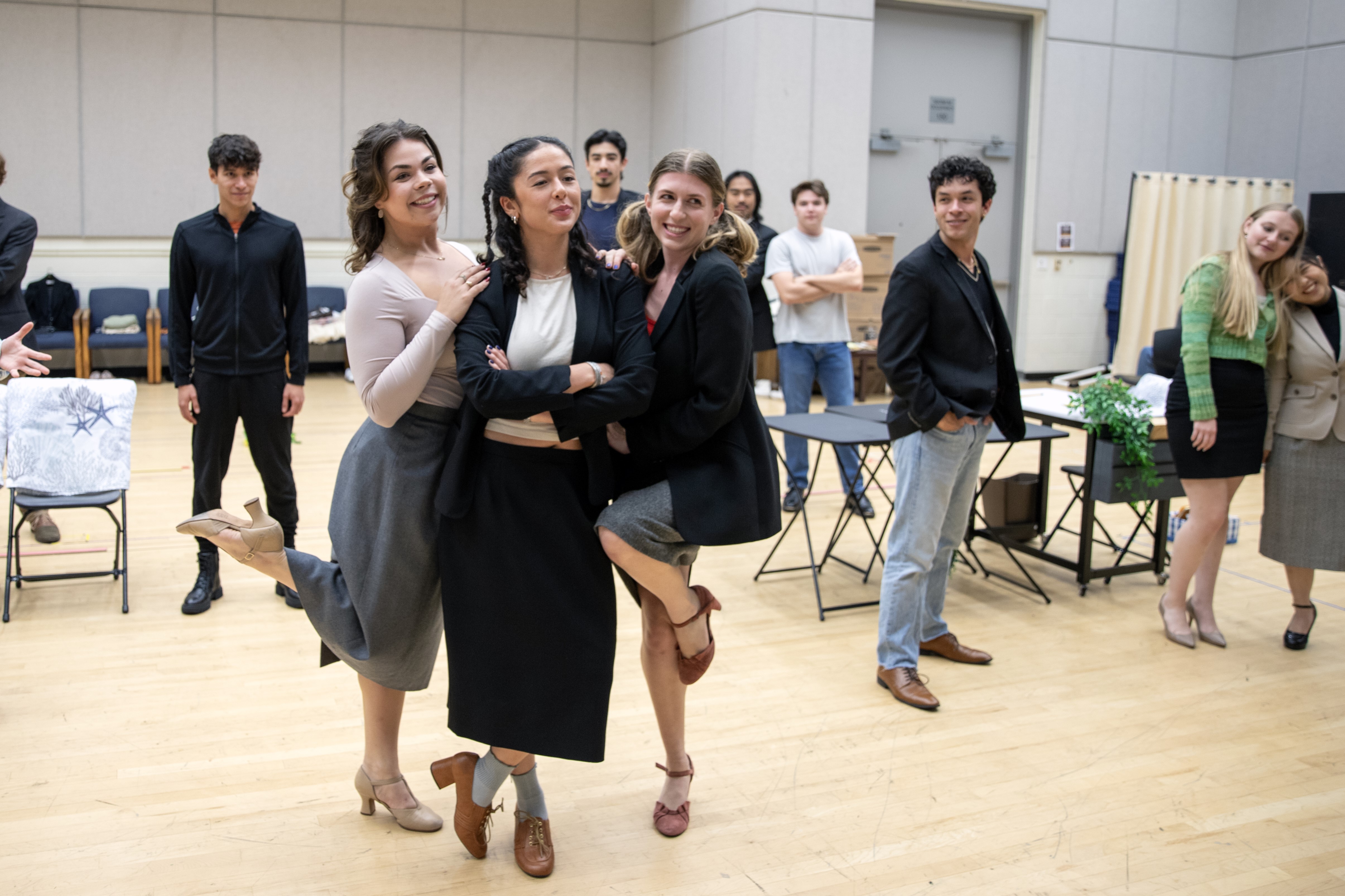 A group of people gathers in a rehearsal studio, with three women at center smiling and posing together. Others stand and watch in the background near scattered chairs and tables, creating a lively, informal atmosphere.
