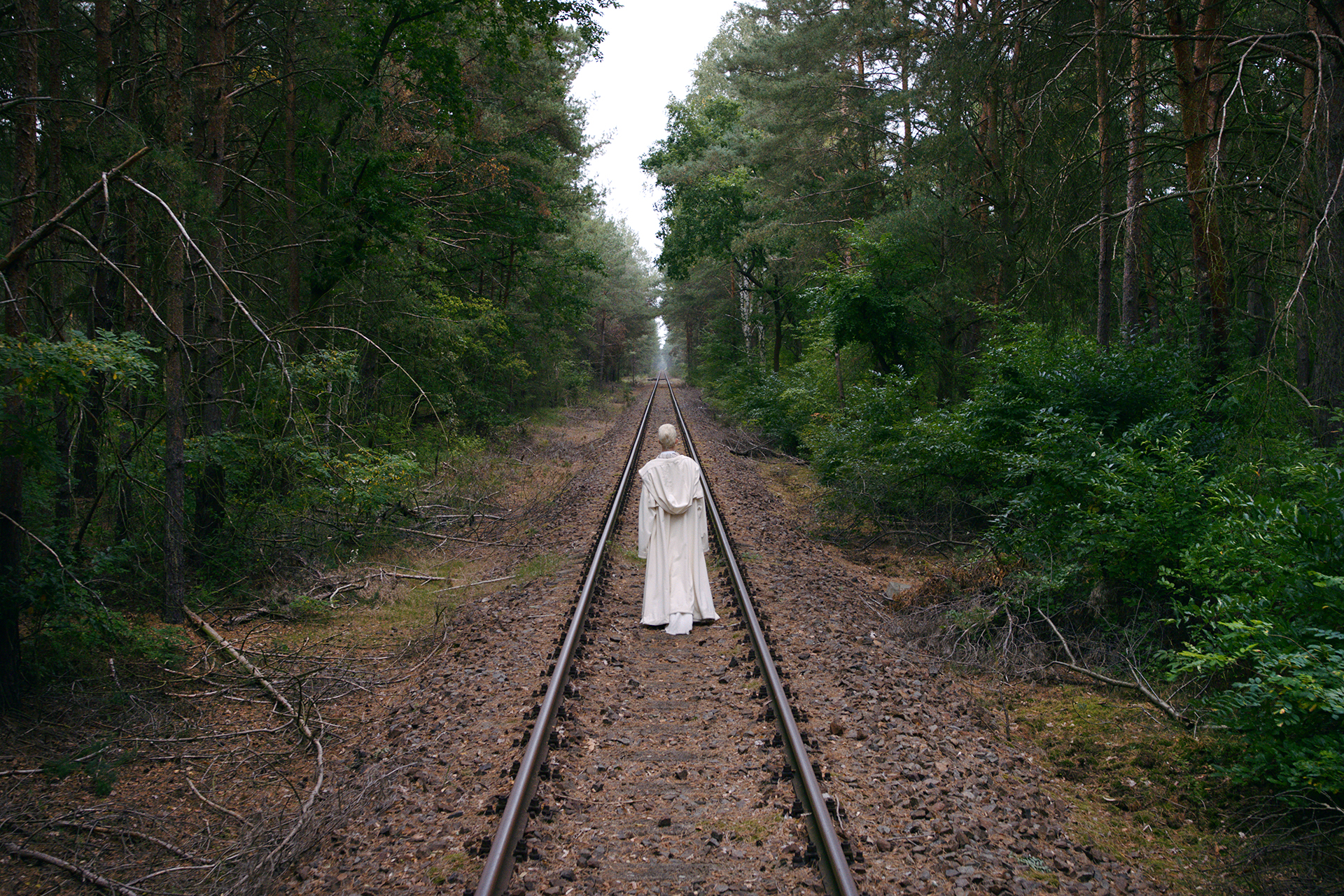 A solitary figure in a long white garment stands centered on railroad tracks stretching straight into the distance. Dense green trees line both sides, forming a symmetrical corridor around the tracks.
