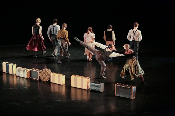 A group of performers moves across a dark stage with vintage-style costumes, while suitcases and trunks are arranged in a line along the floor. One performer balances in a lifted, extended pose as others walk or turn away, creating a layered stage composition.