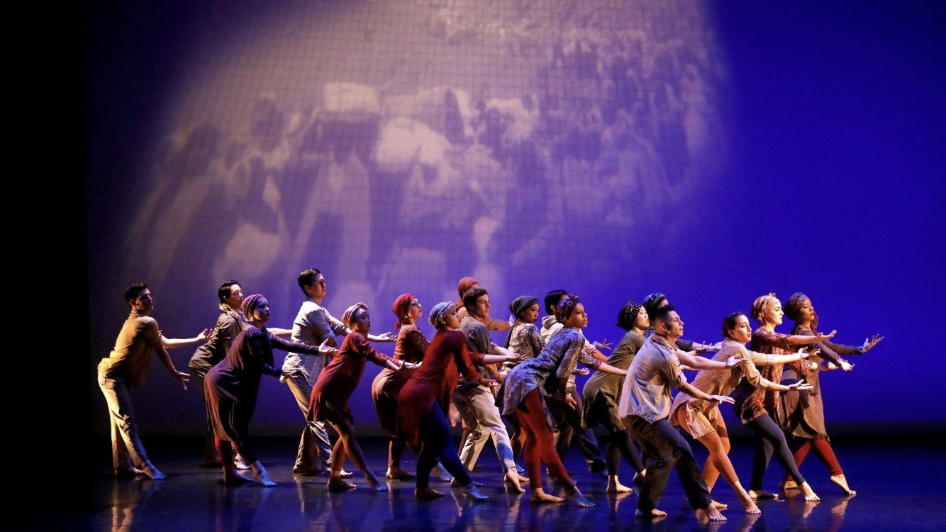 A large group of dancers performs on a stage under deep blue lighting. The performers move in unison, leaning forward with arms extended as if pushing or reaching together. Behind them, a projected black-and-white image fills the backdrop, creating a layered, theatrical atmosphere.
