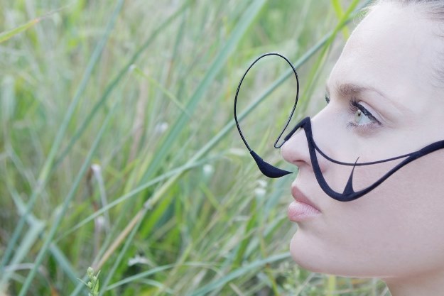 Person in side profile standing among tall green grass, wearing black abstract wire glasses that loop outward in large circular shapes around the eyes.