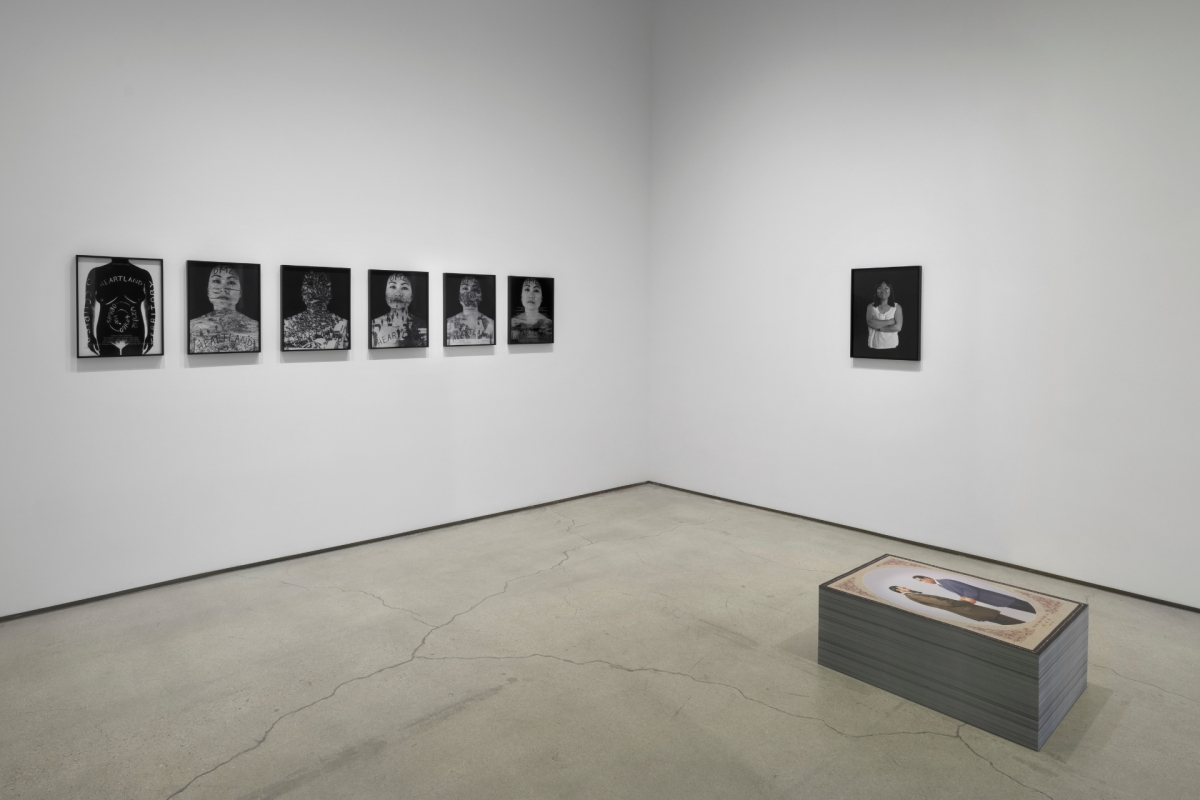 Gallery room with several black-and-white portrait photographs displayed along one wall, a single portrait on an adjacent wall, and a low display case positioned on the floor in the foreground.