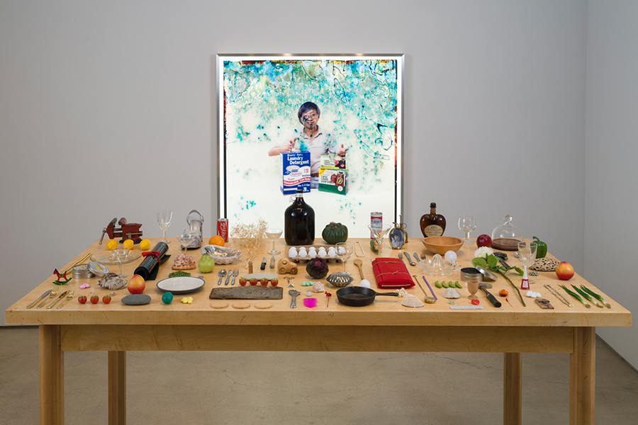 Wooden table displaying a wide assortment of small objects, food items, containers, and tools arranged in rows, with a framed photograph hanging on the wall directly above the table.