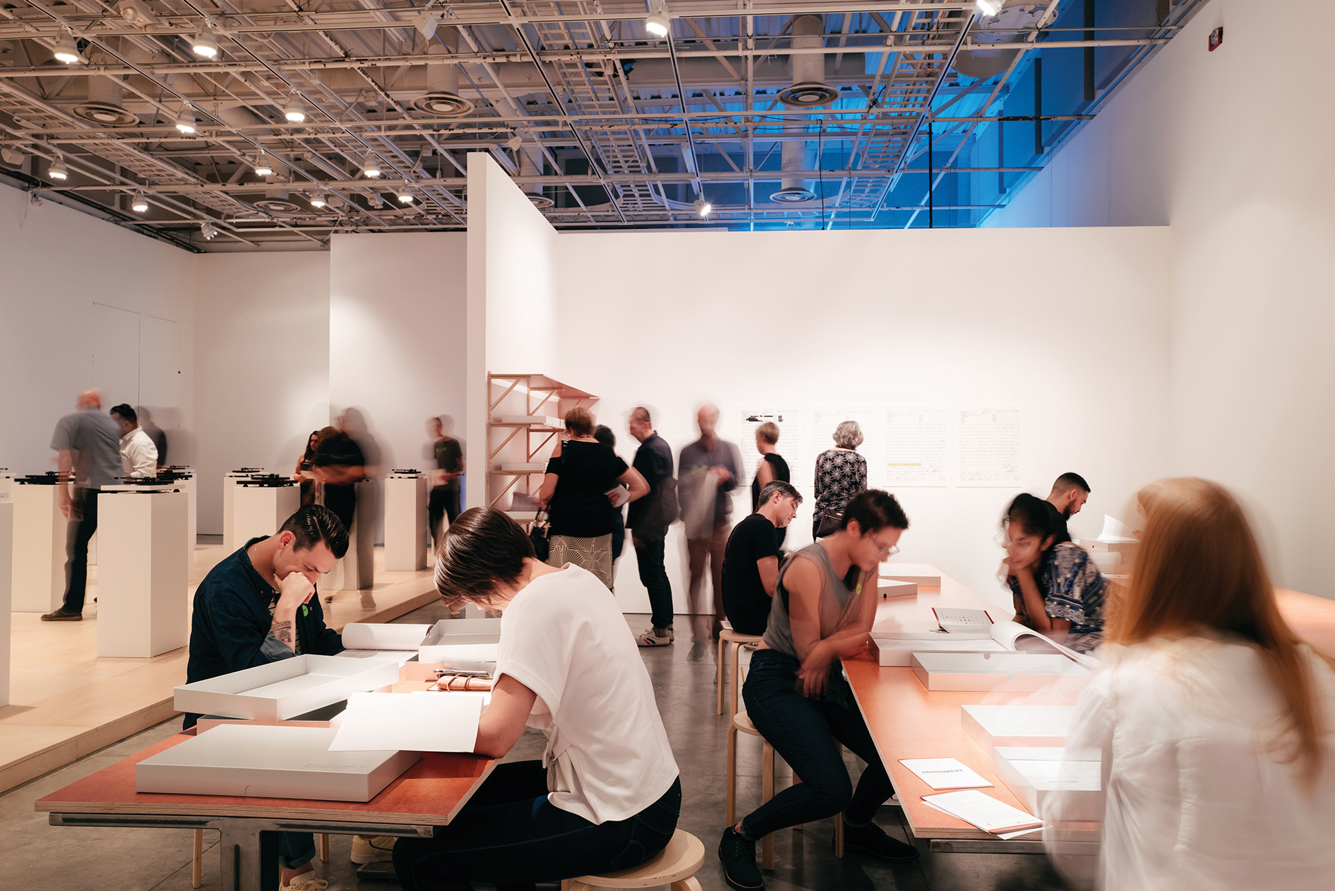 Busy gallery space with visitors viewing displays and sitting at tables examining materials, with exhibition walls and pedestal displays visible under overhead lighting.