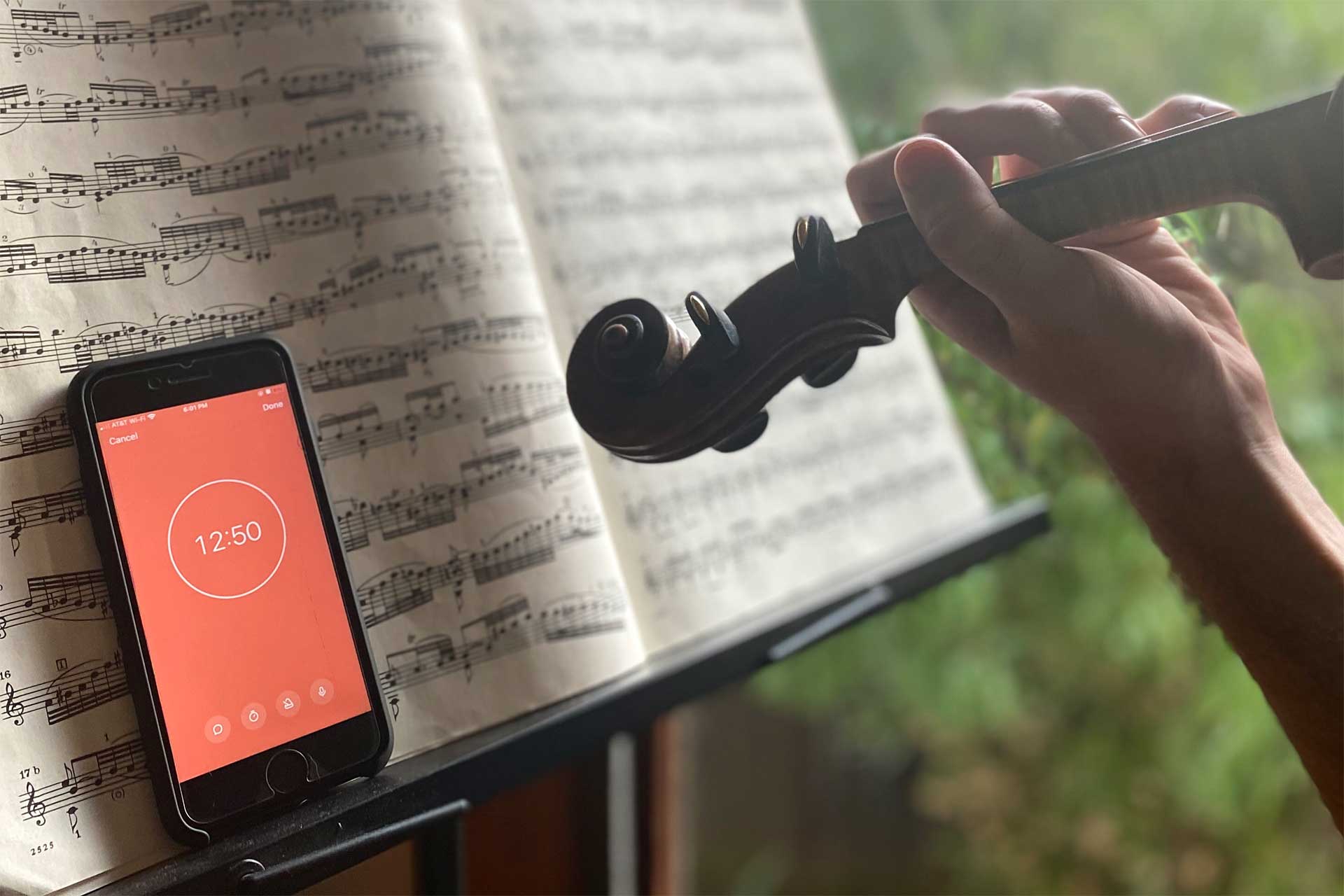 Close-up of a hand holding a violin scroll near a music stand displaying sheet music, with a smartphone on the stand showing a timer set to 12:50.