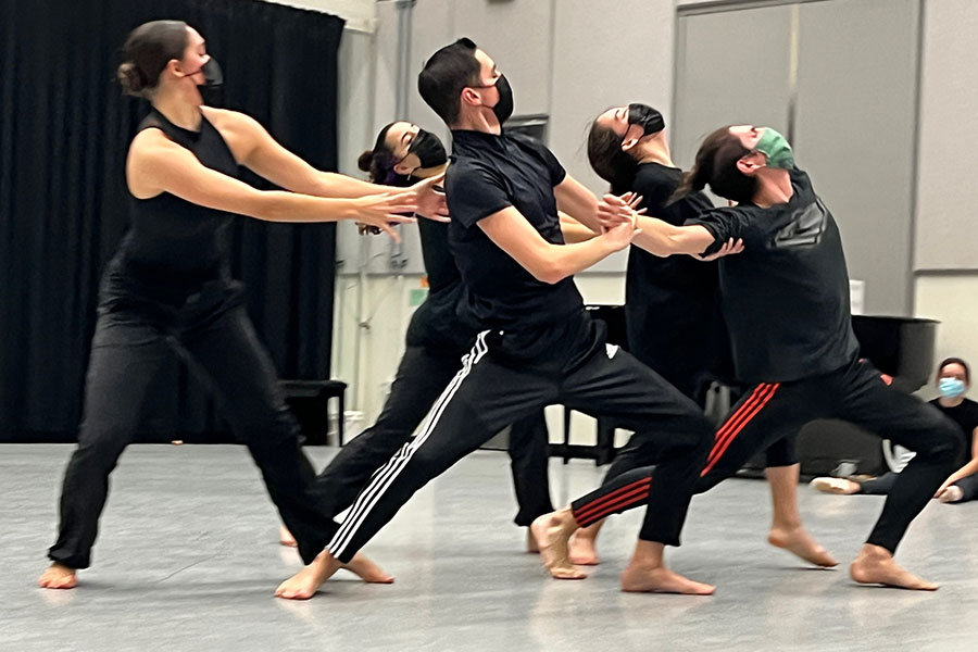 Group of dancers in a studio wearing black rehearsal clothing and face masks, performing a synchronized movement while leaning and reaching together.