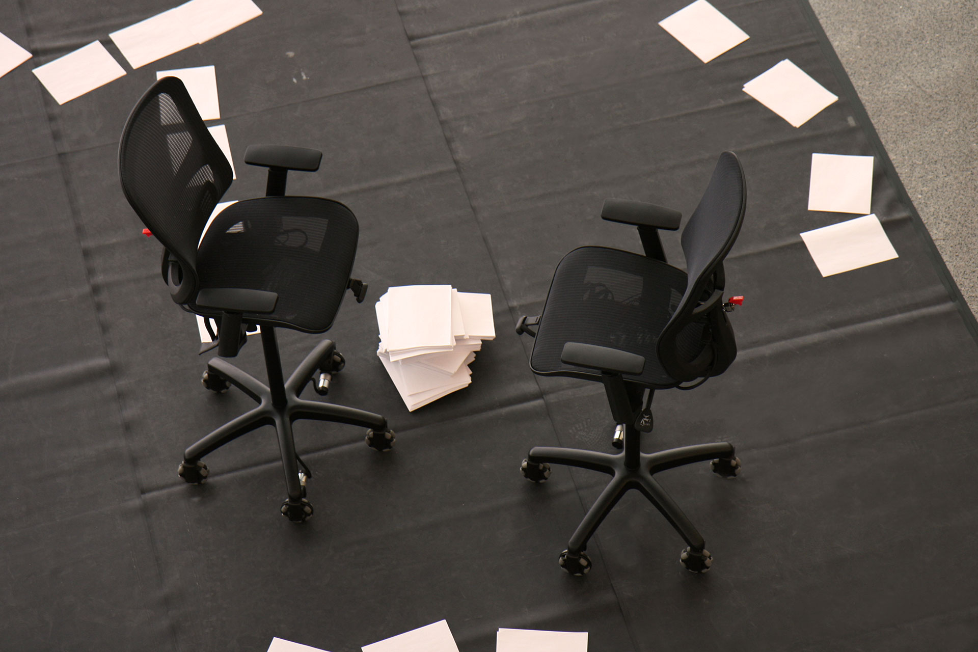 Two black office chairs positioned on a dark floor surface with scattered sheets of white paper surrounding them and a small stack of papers placed between the chairs.