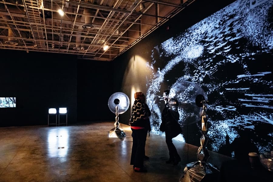 Two people view a large, textured black and white projection art in a dimly lit gallery