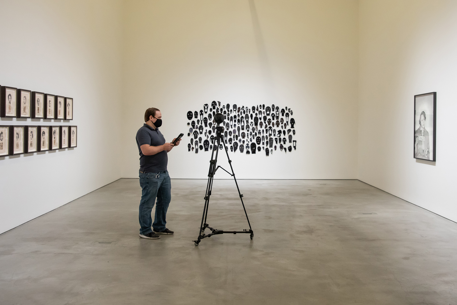 Person standing in a gallery beside a tripod-mounted camera, facing a wall installation composed of many small black silhouette portraits, with additional framed works displayed on the surrounding walls.