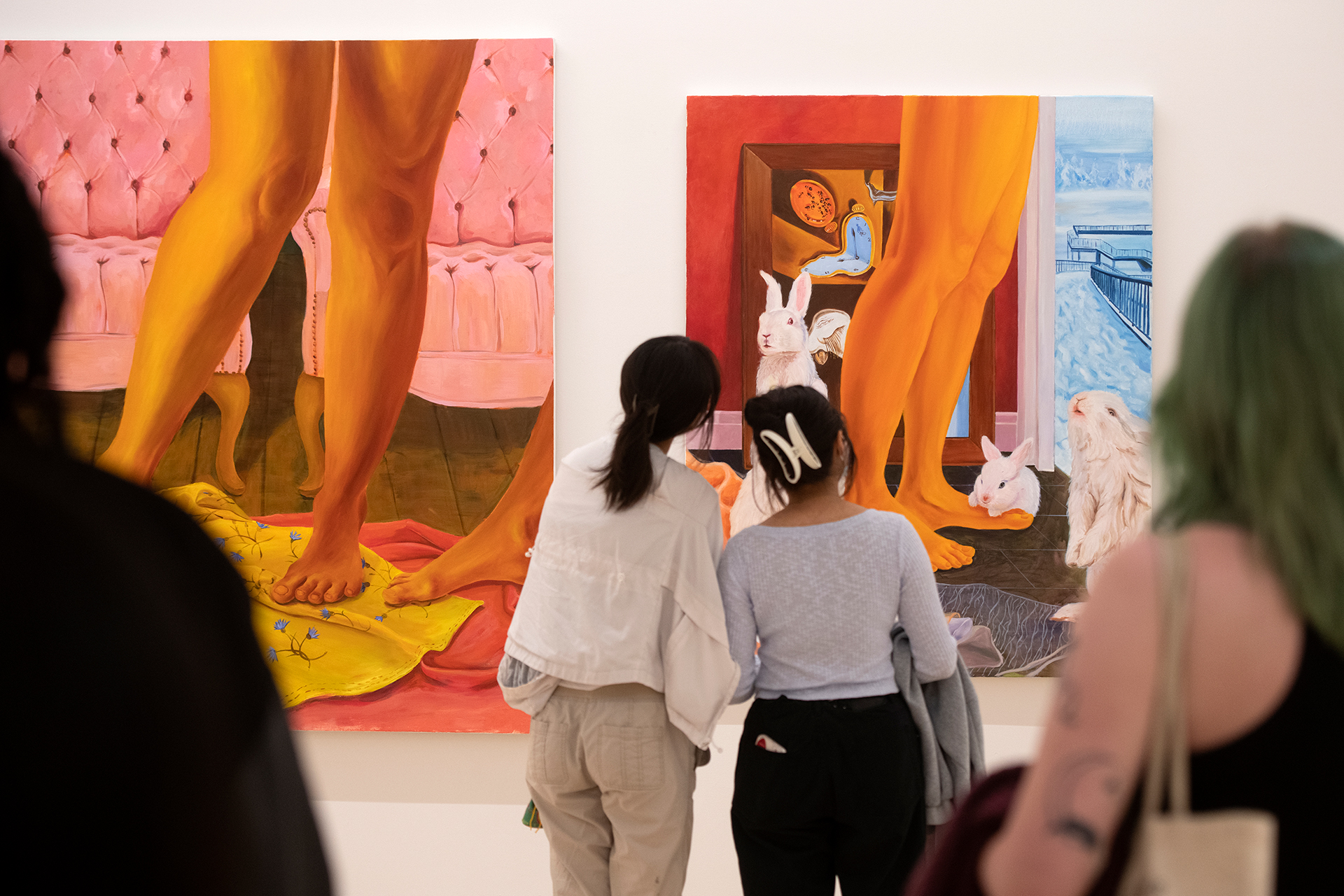 Visitors standing in a gallery looking at two large colorful paintings featuring oversized legs and interior scenes with animals and objects, viewed from behind as they observe the artwork.