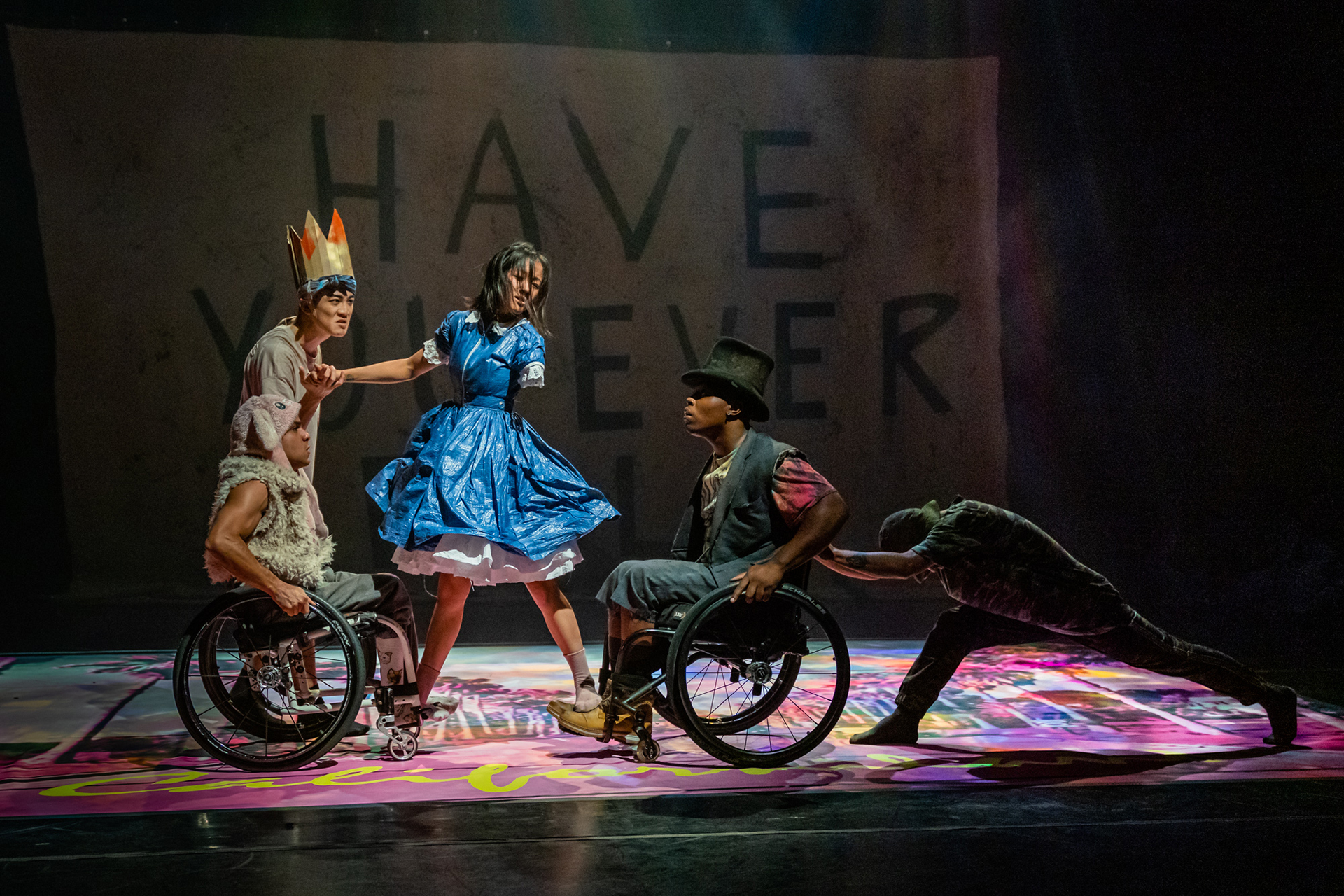 Theatrical stage scene featuring performers in costume, including two individuals in wheelchairs and others interacting dynamically, with colorful lighting and a backdrop displaying large text.